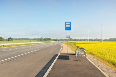 Otobüs durağı ve karayolu (yeni asfalt yolu) yeşil orman çayırı boyunca ve güneş doğarken açan sarı kolza tohumu tarlasında. Sis, yumuşak güneş ışığı. Ulaşım, uzak yerler, tatiller
