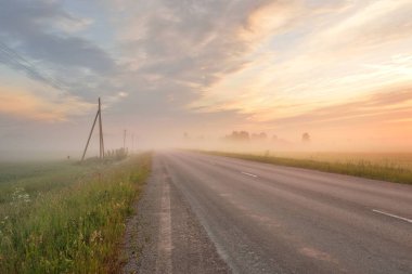 Kırsal karayolu (eski asfalt yolu) gün doğumunda tarla ve ormanın içinden geçer. Yumuşak güneş ışığı, parlayan pembe ve altın bulutlar. Elektrik hattı, trafo direkleri, kablolar. Tatiller, lojistik