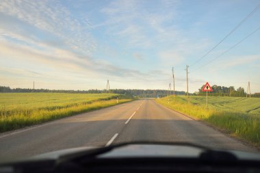 Gündoğumunda tarla ve ormanın içinden (asfalt yol) geçer. Yumuşak güneş ışığı, parlayan bulutlar. Tatiller, lojistik, ücra yerler. Arabadan bak.