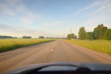 Gündoğumunda tarla ve ormanın içinden (asfalt yol) geçer. Yumuşak güneş ışığı, parlayan bulutlar. Tatiller, lojistik, ücra yerler. Arabadan bak.