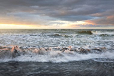 Gün batımında Baltık kıyısı. Yumuşak altın güneş ışığı, dramatik gökyüzü, parlayan bulutlar, dalgalar. Idyllic deniz manzarası. Seyahat yerleri, eko-turizm, doğa, hava durumu, iklim değişikliği. Resimli panoramik manzara