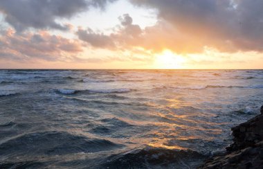 Mavi gökyüzünün altında, parlak pembe ve altın renkli günbatımı bulutlarıyla Baltık deniz kıyısında. Kaya kıyısı, dalgalar. Dalgalar çarpıyor. - Letonya. Doğa, çevre koruma, eko-turizm. Resim gibi bir manzara.