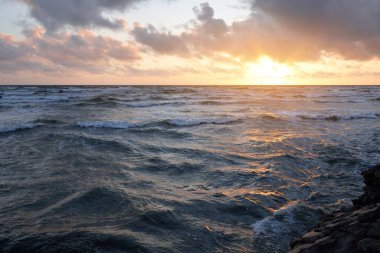 Mavi gökyüzünün altında, parlak pembe ve altın renkli günbatımı bulutlarıyla Baltık deniz kıyısında. Kaya kıyısı, dalgalar. Dalgalar çarpıyor. - Letonya. Doğa, çevre koruma, eko-turizm. Resim gibi bir manzara.