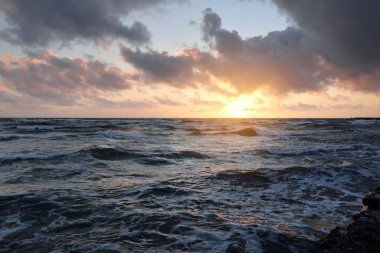 Mavi gökyüzünün altında, parlak pembe ve altın renkli günbatımı bulutlarıyla Baltık deniz kıyısında. Kaya kıyısı, dalgalar. Dalgalar çarpıyor. - Letonya. Doğa, çevre koruma, eko-turizm. Resim gibi bir manzara.