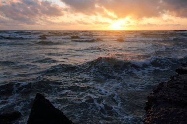 Mavi gökyüzünün altında, parlak pembe ve altın renkli günbatımı bulutlarıyla Baltık deniz kıyısında. Kaya kıyısı, dalgalar. Dalgalar çarpıyor. - Letonya. Doğa, çevre koruma, eko-turizm. Resim gibi bir manzara.