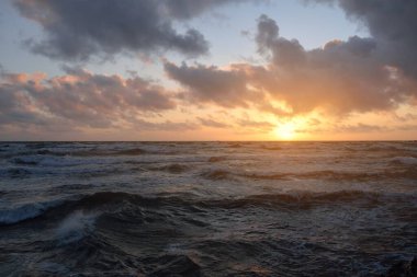 Fırtınadan sonra parlayan pembe ve altın rengi günbatımı bulutlarıyla mavi gökyüzünün altında Baltık Denizi kıyısında. Dalgalar çarpıyor. Doğa, çevre koruma, eko-turizm. Resimli panoramik manzara