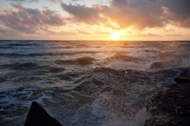 Mavi gökyüzünün altında, parlak pembe ve altın renkli günbatımı bulutlarıyla Baltık deniz kıyısında. Kaya kıyısı, dalgalar. Dalgalar çarpıyor. - Letonya. Doğa, çevre koruma, eko-turizm. Resim gibi bir manzara.