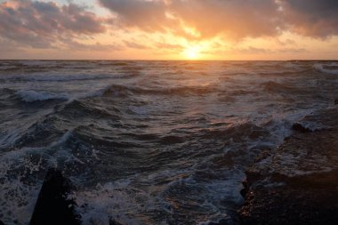 Mavi gökyüzünün altında, parlak pembe ve altın renkli günbatımı bulutlarıyla Baltık deniz kıyısında. Kaya kıyısı, dalgalar. Dalgalar çarpıyor. - Letonya. Doğa, çevre koruma, eko-turizm. Resim gibi bir manzara.
