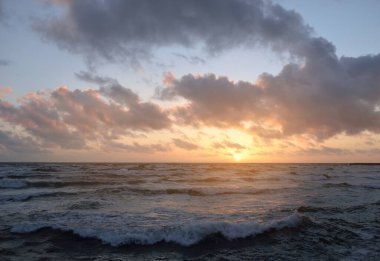 Fırtınadan sonra parlayan pembe ve altın rengi günbatımı bulutlarıyla mavi gökyüzünün altında Baltık Denizi kıyısında. Dalgalar çarpıyor. Doğa, çevre koruma, eko-turizm. Resimli panoramik manzara