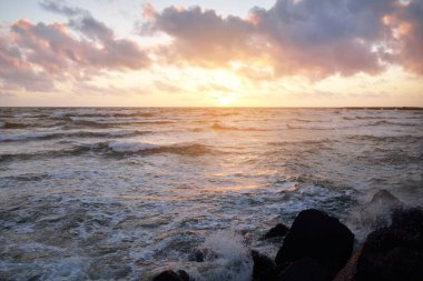 Mavi gökyüzünün altında, parlak pembe ve altın renkli günbatımı bulutlarıyla Baltık deniz kıyısında. Kaya kıyısı, dalgalar. Dalgalar çarpıyor. - Letonya. Doğa, çevre koruma, eko-turizm. Resim gibi bir manzara.