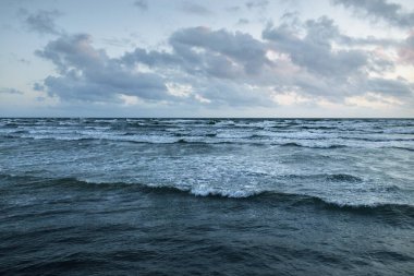 Fırtınadan sonra parlayan günbatımı bulutlarıyla mavi gökyüzünün altında Baltık Denizi kıyısında. Su yüzeyi dokusu, çarpan dalgalar. Doğa, çevre koruma, eko-turizm. Resimli panoramik manzara