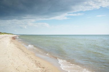 Baltık deniz kıyısı. Açık mavi gökyüzü. Yumuşak güneş ışığı. Kumsal, kum tepeleri, çakıl taşları. Saf doğa, çevre koruma, ekoloji, eko-turizm, yürüyüş, yaz tatili, dinlenme. Panoramik görünüm