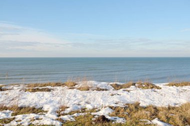 Baltık Denizi 'nin karla kaplı kumlu kıyılardan (kum tepeleri) panoramik görüntüsü. Dramatik gökyüzü, parlayan bulutlar, dalgalar ve su sıçramaları. Deniz Burnu. Sıcak kış havası, iklim değişikliği, doğa. Letonya