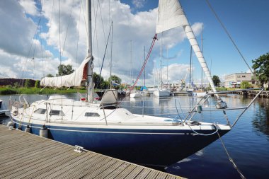 Güneşli bir yaz gününde mavi şalopalı bir yat rıhtıma demirli. Arka planda yelkenliler var. Baltık Denizi, Avrupa. Tatiller, ulaşım, spor, eğlence, turizm, seyahat, balıkçılık, liman hizmeti