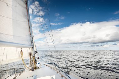Açık bir günde açık denizde yelken açan beyaz yelkenli bir yat. Güverteden pruvaya bir manzara. Kümülüs bulutları. Ulaşım, seyahat, gezi, spor, eğlence, boş zaman aktivitesi, yarış, yelkenli yarışı
