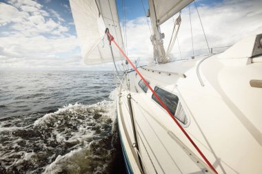 Açık bir günde açık denizde yelken açan beyaz yelkenli bir yat. Güverteden pruvaya bir manzara. Kümülüs bulutları. Ulaşım, seyahat, gezi, spor, eğlence, boş zaman aktivitesi, yarış, yelkenli yarışı