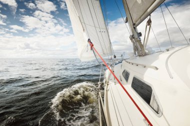 Açık bir günde açık denizde yelken açan beyaz yelkenli bir yat. Güverteden pruvaya bir manzara. Kümülüs bulutları. Ulaşım, seyahat, gezi, spor, eğlence, boş zaman aktivitesi, yarış, yelkenli yarışı