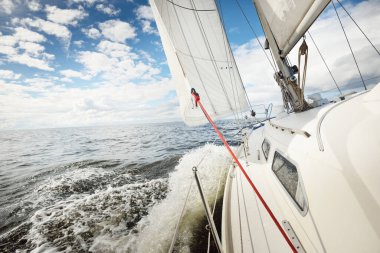 Açık bir günde açık denizde yelken açan beyaz yelkenli bir yat. Güverteden pruvaya bir manzara. Kümülüs bulutları. Ulaşım, seyahat, gezi, spor, eğlence, boş zaman aktivitesi, yarış, yelkenli yarışı