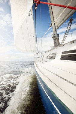 Açık bir günde açık denizde yelken açan beyaz yelkenli bir yat. Güverteden pruvaya bir manzara. Kümülüs bulutları. Ulaşım, seyahat, gezi, spor, eğlence, boş zaman aktivitesi, yarış, yelkenli yarışı