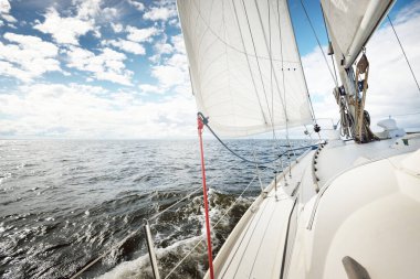 Açık bir günde açık denizde yelken açan beyaz yelkenli bir yat. Güverteden pruvaya bir manzara. Kümülüs bulutları. Ulaşım, seyahat, gezi, spor, eğlence, boş zaman aktivitesi, yarış, yelkenli yarışı