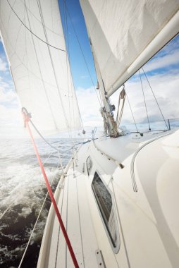 Açık bir günde açık denizde yelken açan beyaz yelkenli bir yat. Güverteden pruvaya bir manzara. Kümülüs bulutları. Ulaşım, seyahat, gezi, spor, eğlence, boş zaman aktivitesi, yarış, yelkenli yarışı