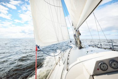 Açık bir günde açık denizde yelken açan beyaz yelkenli bir yat. Güverteden pruvaya bir manzara. Kümülüs bulutları. Ulaşım, seyahat, gezi, spor, eğlence, boş zaman aktivitesi, yarış, yelkenli yarışı