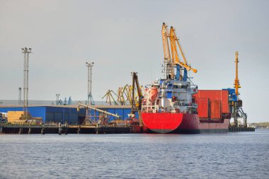 Liman terminalinde büyük bir kargo gemisi yükleniyor, arka planda vinçler var. Yük taşımacılığı, lojistik, küresel iletişim, ekonomi, ticaret, sanayi, özel ekipman