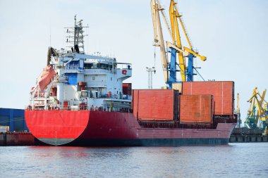 Liman terminalinde büyük bir kargo gemisi yükleniyor, arka planda vinçler var. Yük taşımacılığı, lojistik, küresel iletişim, ekonomi, ticaret, sanayi, özel ekipman