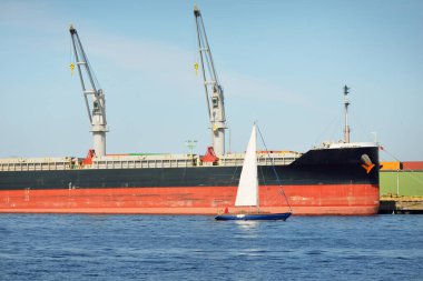 Baltık Denizi 'nde yelkenli bir yat. Kargo gemisi arka plandaki iskele terminaline yükleniyor. Ulaşım, seyahat, gezinti, spor, eğlence, eğlence etkinliği, iş, ekonomi, sanayi