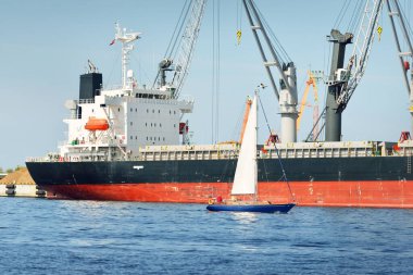 Baltık Denizi 'nde yelkenli bir yat. Kargo gemisi arka plandaki iskele terminaline yükleniyor. Ulaşım, seyahat, gezinti, spor, eğlence, eğlence etkinliği, iş, ekonomi, sanayi