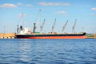 Liman terminalinde büyük bir kargo gemisi yükleniyor, arka planda vinçler var. Yük taşımacılığı, lojistik, küresel iletişim, ekonomi, ticaret, sanayi, özel ekipman