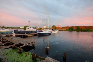 Eski römorkör ve mavi yelkenli yelkenli gün batımında yat limanındaki bir rıhtıma demirlemiş. Dramatik bir gökyüzü. Tatiller, ulaşım, spor, eğlence, turizm, seyahat, balıkçılık, liman hizmeti