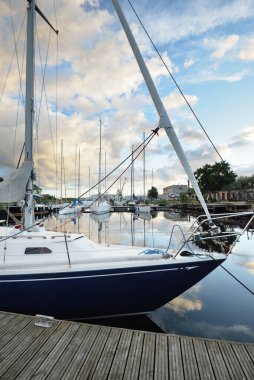 Gün batımında yat limanındaki bir rıhtıma demirlenmiş mavi yelkenli. Dramatik gökyüzü, parlayan altın bulutlar. Tatiller, ulaşım, spor, eğlence, turizm, seyahat, liman