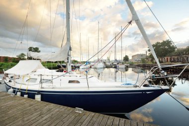 Gün batımında yat limanındaki bir rıhtıma demirlenmiş mavi yelkenli. Dramatik gökyüzü, parlayan altın bulutlar. Tatiller, ulaşım, spor, eğlence, turizm, seyahat, liman