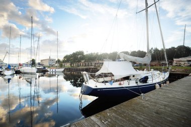 Gün batımında yat limanındaki bir rıhtıma demirlenmiş mavi yelkenli. Dramatik gökyüzü, parlayan altın bulutlar. Tatiller, ulaşım, spor, eğlence, turizm, seyahat, liman