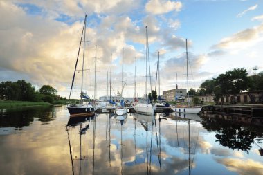 Günbatımında yat limanında demirlemiş zarif ve modern yelkenliler. Dramatik gökyüzü, parlayan altın bulutlar. Tatiller, ulaşım, spor, eğlence, turizm, seyahat, liman