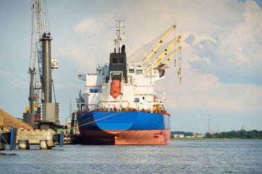 Liman terminalinde büyük bir kargo gemisi yükleniyor, arka planda vinçler var. Yük taşımacılığı, deniz taşımacılığı, lojistik, küresel iletişim, dünya çapında taşımacılık, ekonomi, ticaret, sanayi