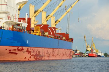 Liman terminalinde büyük bir kargo gemisi yükleniyor, arka planda vinçler var. Yük taşımacılığı, deniz taşımacılığı, lojistik, küresel iletişim, dünya çapında taşımacılık, ekonomi, ticaret, sanayi