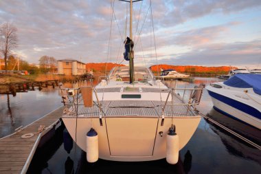 Günbatımında yat limanındaki bir rıhtıma demirlenmiş ahşap meşe güverteli modern yelkenli. Dramatik gökyüzü, parlayan altın bulutlar. Tatiller, ulaşım, spor, eğlence, turizm, seyahat, liman