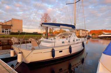 Günbatımında yat limanındaki bir rıhtıma demirlenmiş ahşap meşe güverteli modern yelkenli. Dramatik gökyüzü, parlayan altın bulutlar. Tatiller, ulaşım, spor, eğlence, turizm, seyahat, liman