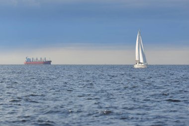 Baltık Denizi 'nde yelkenli bir yat, arkada kargo gemisi. Fırtınadan sonra açık gökyüzü, yumuşak güneş ışığı. Ulaşım, seyahat, gezi, spor, eğlence, boş zaman aktivitesi, yelkenli yarışı