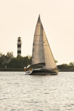 Baltık Denizi 'nde yelkenli bir yat. Deniz fenerine yakın çekim. Fırtınadan sonra gün batımı gökyüzü açık. Ulaşım, seyahat, gezi, spor, eğlence, boş zaman aktivitesi, yarış, yelkenli yarışı