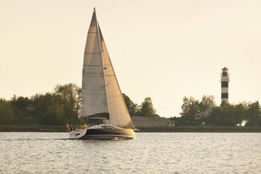 Baltık Denizi 'nde beyaz yelkenli yelkenli. Deniz fenerine yakın çekim. Fırtınadan sonra gün batımı gökyüzü açık. Ulaşım, seyahat, gezi, spor, eğlence, boş zaman aktivitesi, yarış, yelkenli yarışı