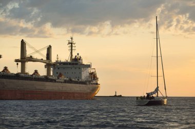 Büyük kargo gemisi (büyük yük gemisi, 179.99 metre uzunluğunda) ve günbatımında Baltık Denizi 'nde yelkenli yelkenli. Yük taşımacılığı, küresel iletişim, ekonomi, seyahat, eğlence. spor