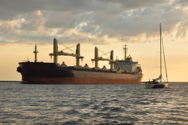 Büyük kargo gemisi (büyük yük gemisi, 179.99 metre uzunluğunda) ve günbatımında Baltık Denizi 'nde yelkenli yelkenli. Yük taşımacılığı, küresel iletişim, ekonomi, seyahat, eğlence. spor