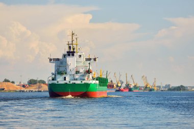 Büyük yeşil yük gemisi gün batımında kargo terminaline varacak. Arka planda gemiler ve vinçler var. Dramatik bulut manzarası. Yük taşımacılığı, küresel iletişim, endüstri, ekonomi, lojistik