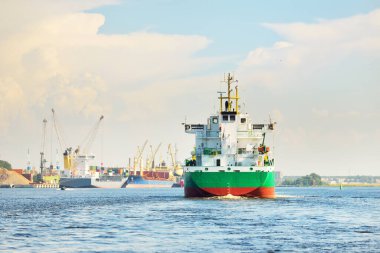 Büyük yeşil yük gemisi gün batımında kargo terminaline varacak. Arka planda gemiler ve vinçler var. Dramatik bulut manzarası. Yük taşımacılığı, küresel iletişim, endüstri, ekonomi, lojistik