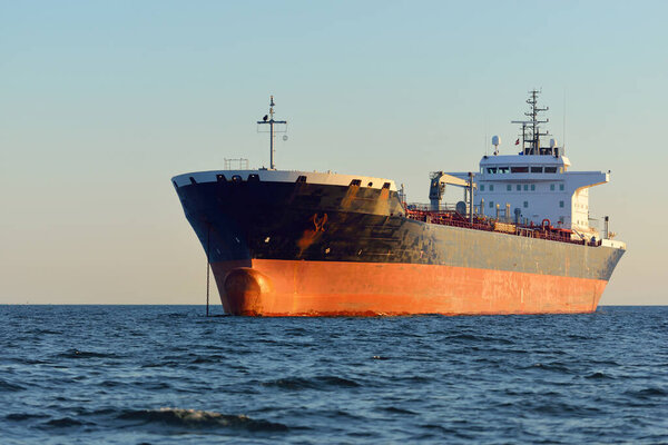 Large cargo ship (chemical tanker, 184 meters length) sailing in an open sea at sunset. Golden sunlight. Freight transportation, fuel, power generation, nautical vessel, logistics, economy, industry