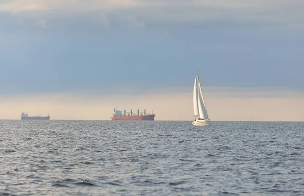 白いスループは バルト海でヨットを操縦し バックグラウンドで貨物船を操縦しました 嵐の後に澄んだ空 柔らかい日差し クルーズ スポーツ レクリエーション レジャー活動 レガッタ — ストック写真