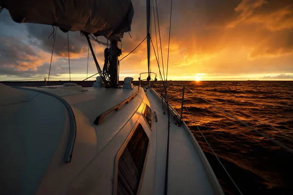 White yacht sailing in an open sea at sunset. A view from the deck to the bow, mast, sails. Epic cloudscape. Dramatic sky with glowing golden clouds after the storm. Racing, sport, leisure activity